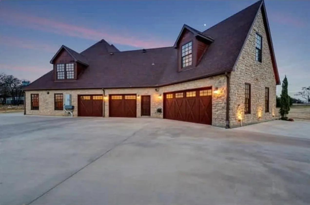 Side of a limestone custom home with cedar carriage garage doors, dormers and a wide concrete drive at dusk